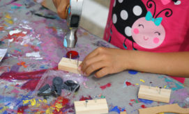 A child's hand uses a hammer to drive nails into small wooden blocks on a colorful, paint-splattered table. A packet of screws and toy parts lies nearby.