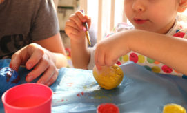 Children paint colorful rocks at a table, using brushes and vibrant paints, including red, blue, and yellow. They sit close together, focused and engaged in the creative activity.