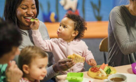 A child wearing a pink hoodie feeds a smiling woman a cucumber slice at a table. Surrounding them are other adults and kids, with visible plates of assorted vegetables.