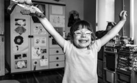 A child with glasses, smiling widely and holding up a toy, stands in a room decorated with drawings and shelves filled with toys.