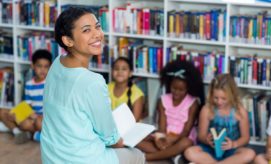 A smiling woman holding a book sits in front of children, who are also holding books, in a library with shelves full of colorful books.