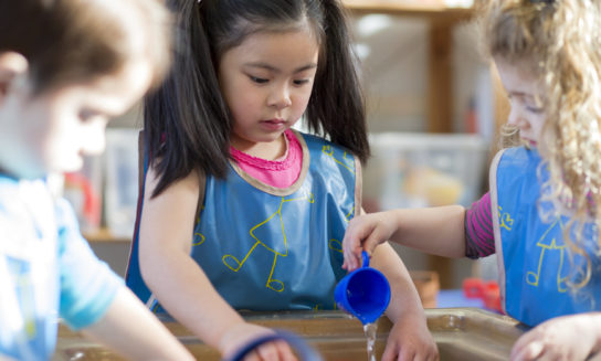 Children wearing blue smocks play with water at a sensory table. One girl pours water from a cup while others observe and participate. The setting is a bright classroom.