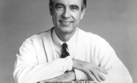 A smiling person in a sweater and tie is resting their arms on a small model labeled "NEIGHBORHOOD TROLLEY" against a plain backdrop.