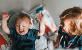 A smiling baby raises its arms while sitting on a soft bed, next to a slightly older child adorned with a necklace, inside a cozy room with patterned pillows.