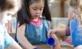 Children wear blue aprons and engage in water play; one pours water from a blue cup, while others watch and participate, in a brightly lit classroom setting.