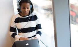 A person wearing headphones is using a laptop with an Apple logo, seated against a large window in a bright indoor space.