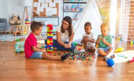 Children and a woman sit on a wooden floor building with colorful plastic blocks. A bright room surrounds them, with a tent, shelves, and a bulletin board in the background.