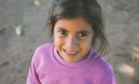 A young girl, wearing a pink sweater, smiles warmly while standing on a dirt-covered ground, surrounded by scattered leaves in an outdoor setting.