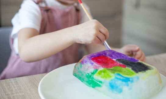 A young girl wearing pink overalls uses a paintbrush to paint with watercolors on a large block of ice.