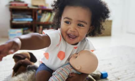 A smiling toddler girl playing with a doll in her classroom, exploring pretend play and emerging social-emotional skills.