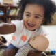 A smiling toddler girl playing with a doll in her classroom, exploring pretend play and emerging social-emotional skills.