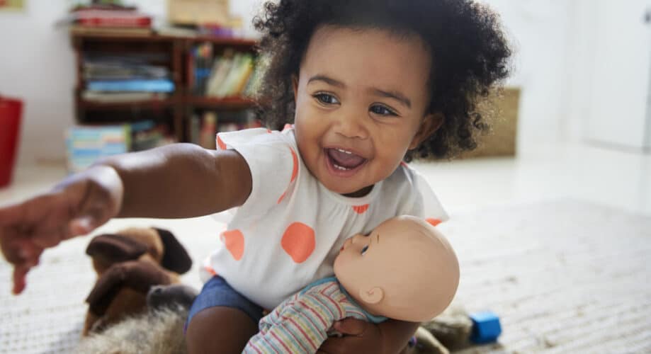 A smiling toddler girl playing with a doll in her classroom, exploring pretend play and emerging social-emotional skills.