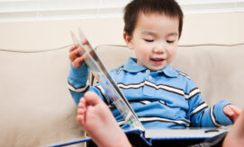 A toddler boy in a blue striped shirt sits on a couch with feet up, engaged in a picture book—demonstrating early literacy exploration.