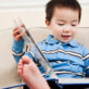 A toddler boy in a blue striped shirt sits on a couch with feet up, engaged in a picture book—demonstrating early literacy exploration.