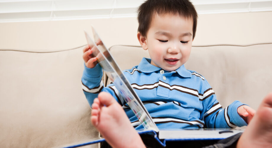 A toddler boy in a blue striped shirt sits on a couch with feet up, engaged in a picture book—demonstrating early literacy exploration.