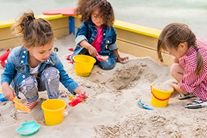 Three preschool girls playing in sand box with buckets and shovels.