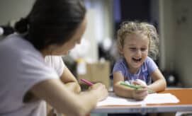 A preschool-aged girl laughs while working on an art activity at a small table with a care provider.