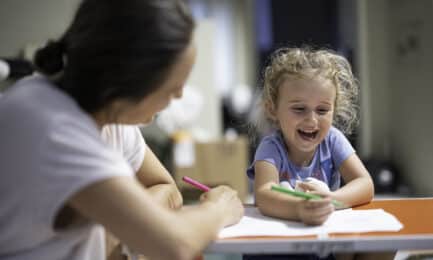 A preschool-aged girl laughs while working on an art activity at a small table with a care provider.