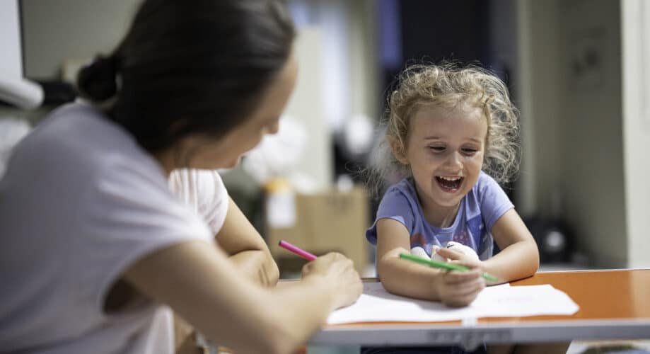 A preschool-aged girl laughs while working on an art activity at a small table with a care provider.