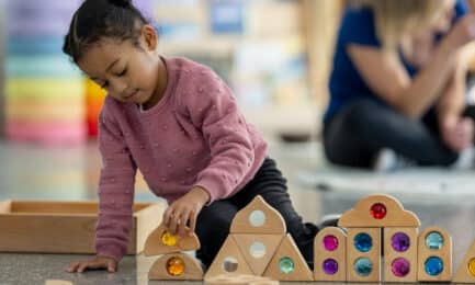 Young girl in pink sweater builds structures with colorful gem blocks in early learning classroom.