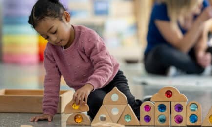 Young girl in pink sweater builds structures with colorful gem blocks in early learning classroom.