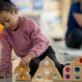Young girl in pink sweater builds structures with colorful gem blocks in early learning classroom.