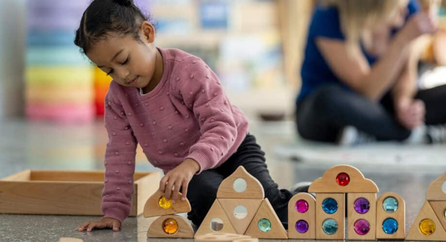 Young girl in pink sweater builds structures with colorful gem blocks in early learning classroom.