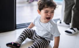 Toddler boy with curly hair, sitting on floor crying while caregiver stands beside him to assist and provide comfort.