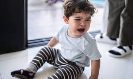 Toddler boy with curly hair, sitting on floor crying while caregiver stands beside him to assist and provide comfort.