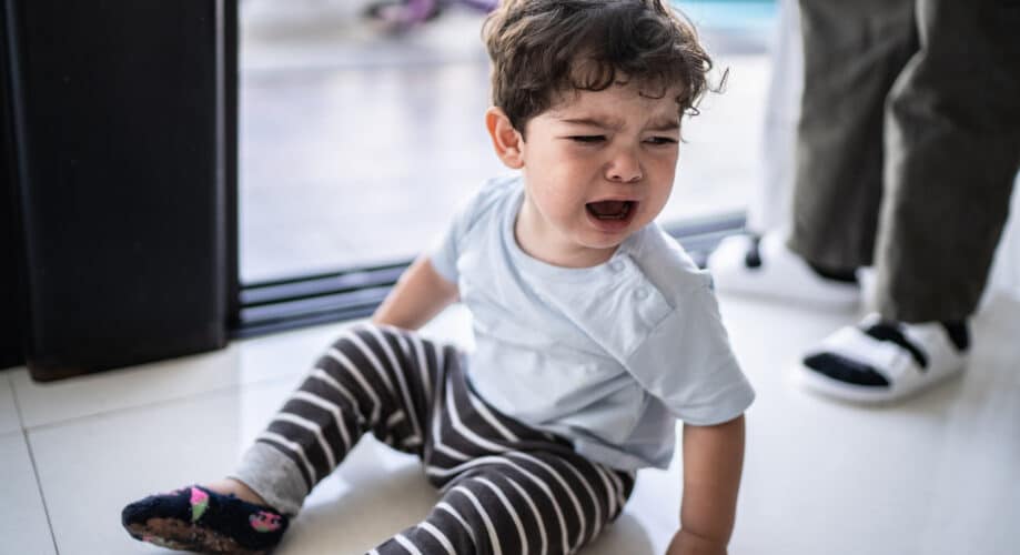 Toddler boy with curly hair, sitting on floor crying while caregiver stands beside him to assist and provide comfort.