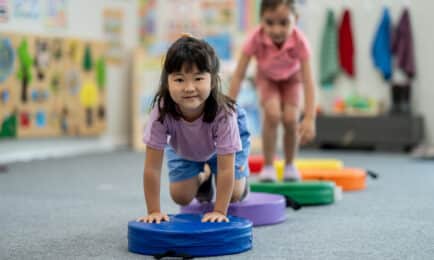 Two preschool-aged girls crawling over an indoor obstacle course to build motor skills through play.