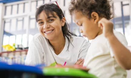 Preschool teacher smiles while sitting next to toddler at art table in early learning classroom.