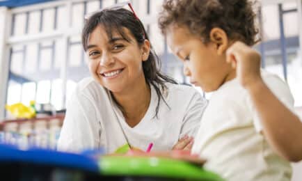 Preschool teacher smiles while sitting next to toddler at art table in early learning classroom.