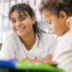 Preschool teacher smiles while sitting next to toddler at art table in early learning classroom.