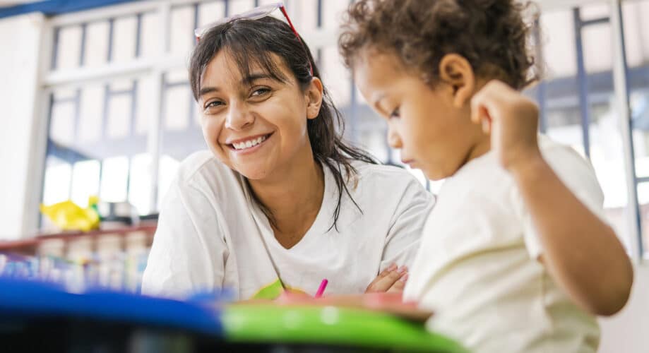 Preschool teacher smiles while sitting next to toddler at art table in early learning classroom.
