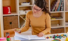An early childhood educator sits at a table in her classroom and reviews paperwork, while surrounded by children’s toys and materials.