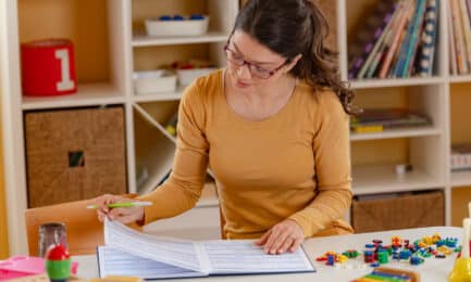 An early childhood educator sits at a table in her classroom and reviews paperwork, while surrounded by children’s toys and materials.