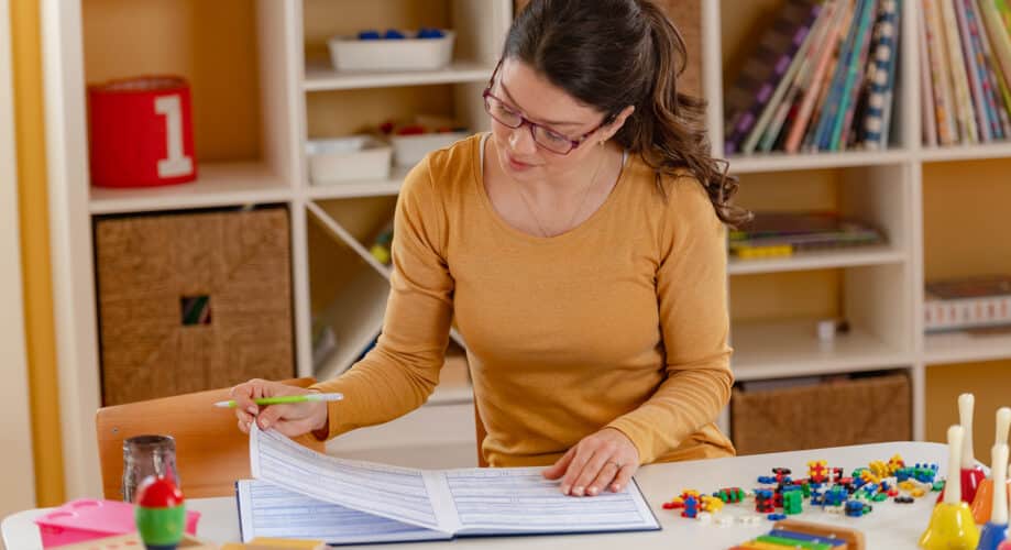 An early childhood educator sits at a table in her classroom and reviews paperwork, while surrounded by children’s toys and materials.