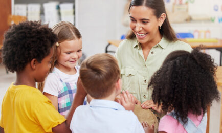 Group of preschool children, smiling, as they join around their educator and join her in playing a musical instrument.