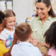 Group of preschool children, smiling, as they join around their educator and join her in playing a musical instrument.