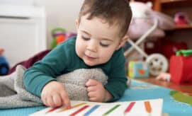 A young child lies on a colorful play mat, pointing to a picture book with colored pencils on the page, building early literacy skills.