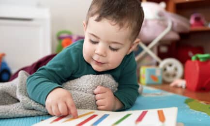 A young child lies on a colorful play mat, pointing to a picture book with colored pencils on the page, building early literacy skills.