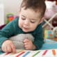 A young child lies on a colorful play mat, pointing to a picture book with colored pencils on the page, building early literacy skills.