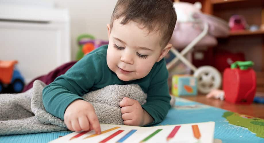 A young child lies on a colorful play mat, pointing to a picture book with colored pencils on the page, building early literacy skills.
