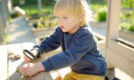 Preschool-aged child on bench outside while looking at rocks with a magnifying glass.