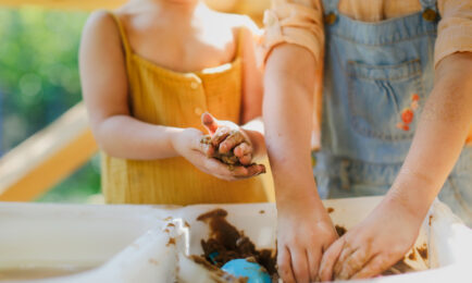 IMAGE ALT TEXT: Two young children enjoying sensory play outdoors, mixing water and sand in a large tub.