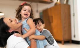 An early learning teacher gives two toddler children a hug while smiling and laughing.