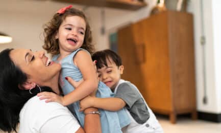 An early learning teacher gives two toddler children a hug while smiling and laughing.