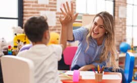 A preschool child sits at a table with his teacher as the two high-five.