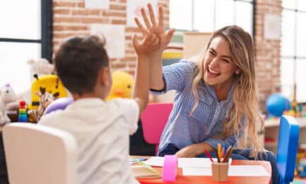 A preschool child sits at a table with his teacher as the two high-five.